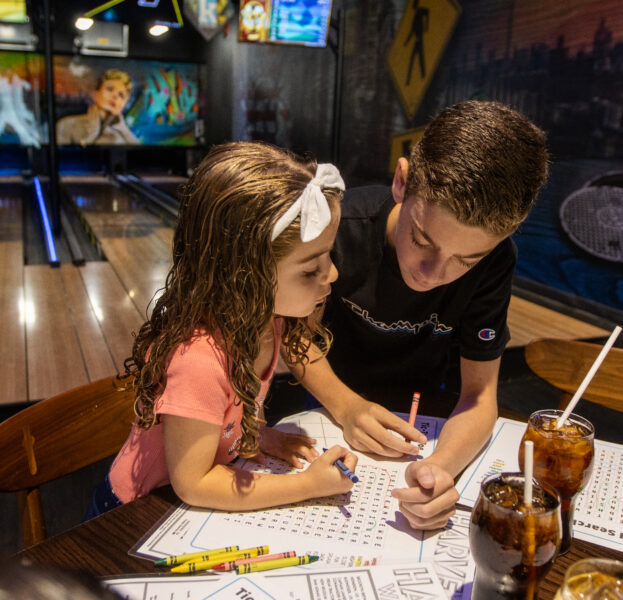 Two kids sit at a table in a bowling alley, working together on a puzzle with crayons. Drinks and menus are on the table beside them.