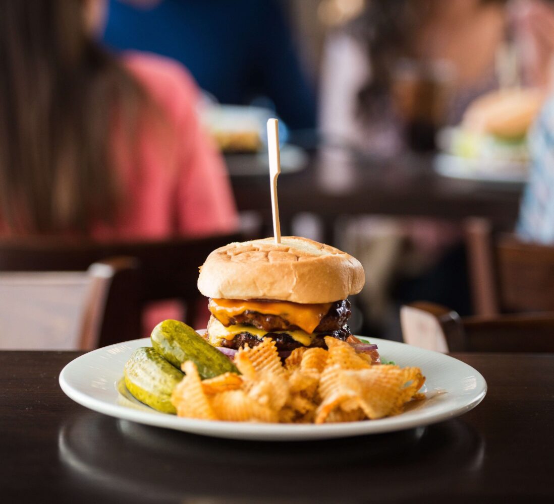 A cheeseburger with pickles and crinkle-cut chips on a plate, with people sitting at a restaurant table in the background.