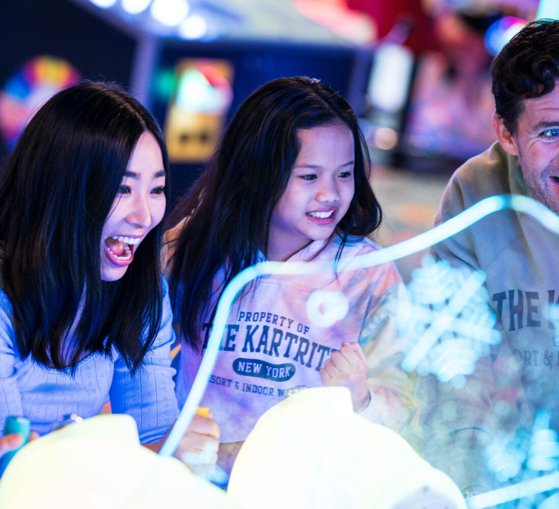 Three people, two adults and a child, excitedly play an arcade game together, illuminated by colorful lights in a lively indoor setting.