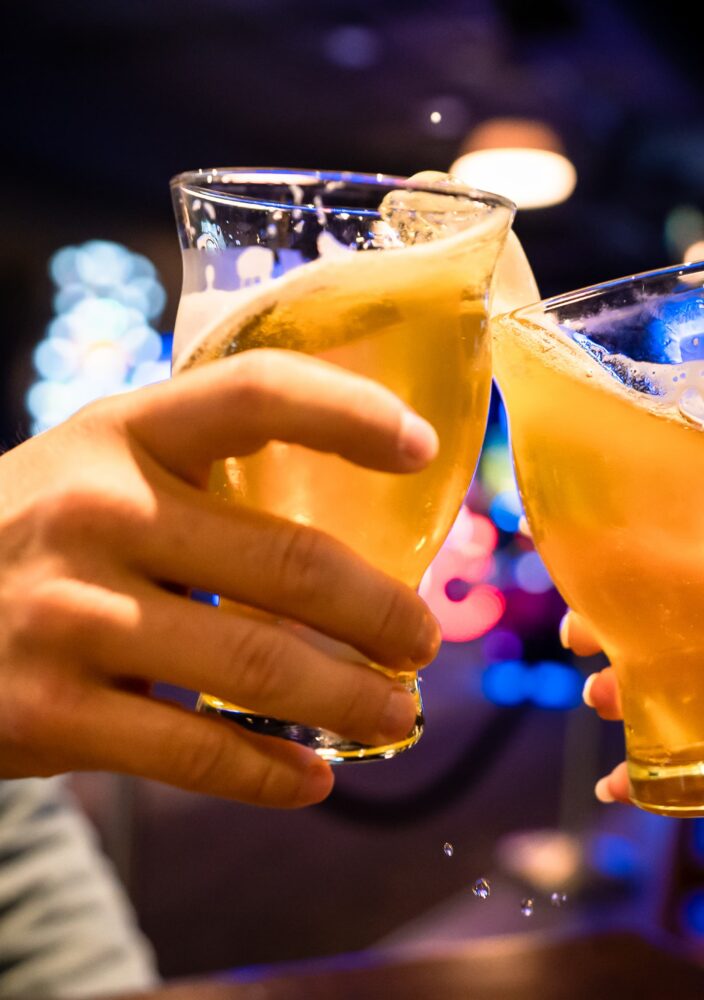 Two people clink glasses of beer together in a dimly lit bar, with blurred colorful lights in the background.