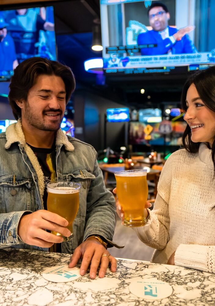 Man and woman smiling, holding pints of beer, sitting at a bar with sports games on TVs in the background.