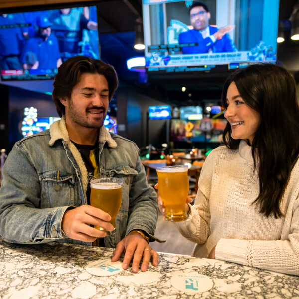 A man and woman smiling and clinking glasses of beer at a bar with TVs and colorful lights in the background.