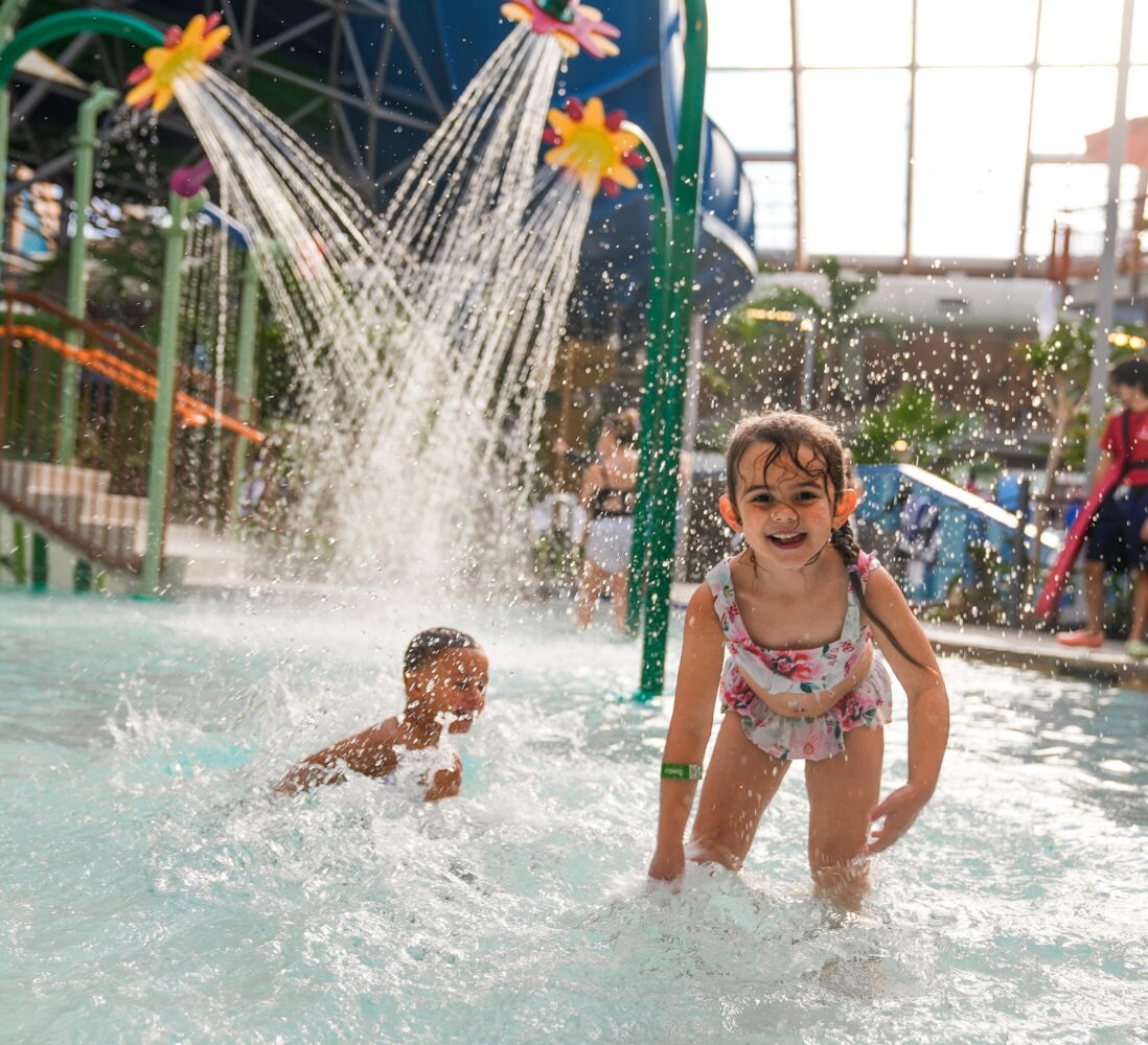 Two kids play in shallow water under flower-shaped sprinklers in an indoor water park, smiling and splashing happily.