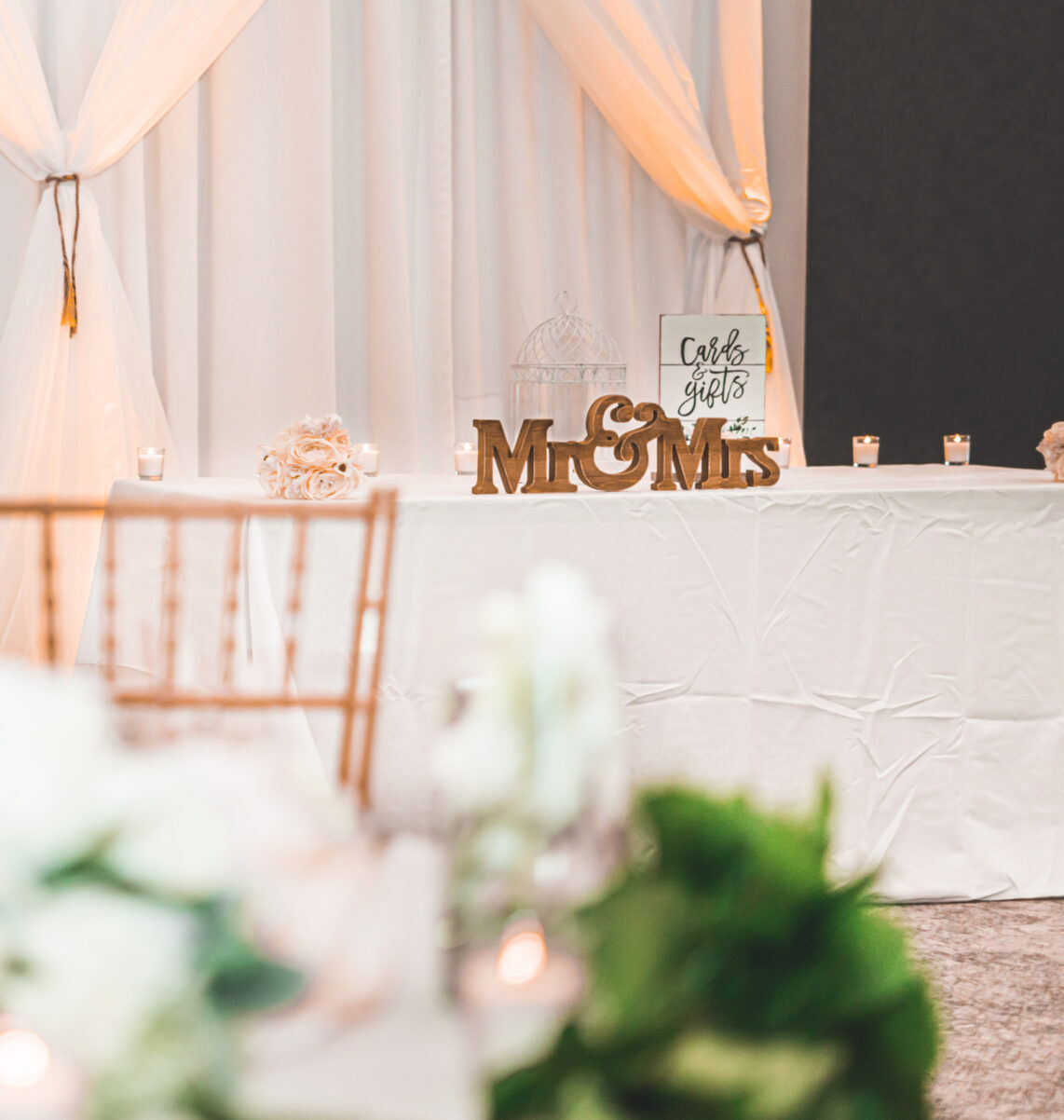 Elegant wedding reception setup with white drapes, gold chairs, floral centerpieces, and a Mr & Mrs sign on a decorated table.