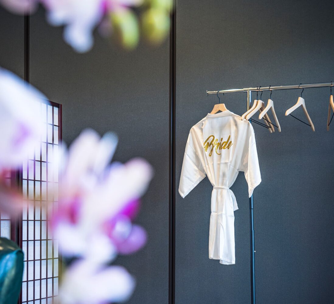 A white robe with Bride in gold hangs on a rack by empty hangers. Blurred pink flowers are in the foreground.