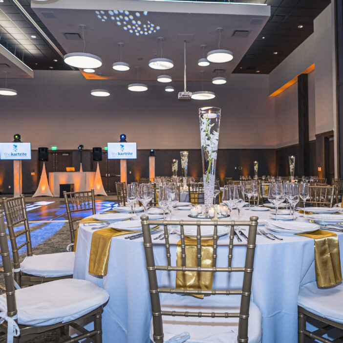 Elegant banquet hall with round tables, gold chairs, tall centerpieces, and screens displaying a logo, set for a formal event.