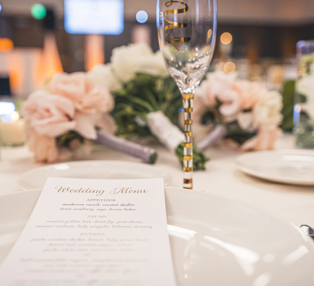 Close-up of a wedding table setting with a menu on a plate, champagne glass, flowers, and candles in the background.
