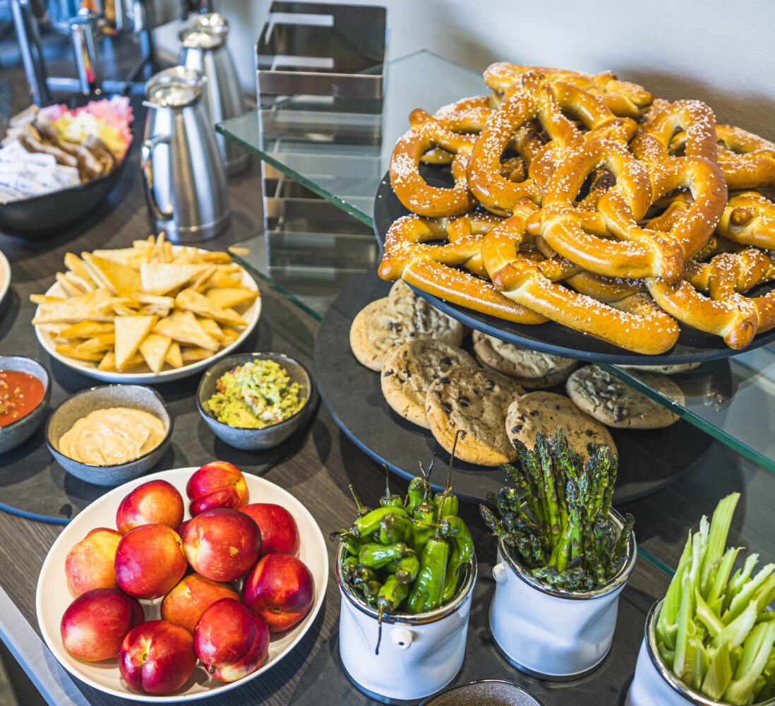 Assorted snacks on a table, including pretzels, cookies, chips, dips, oranges, nectarines, grilled vegetables, and drinks in silver pitchers.