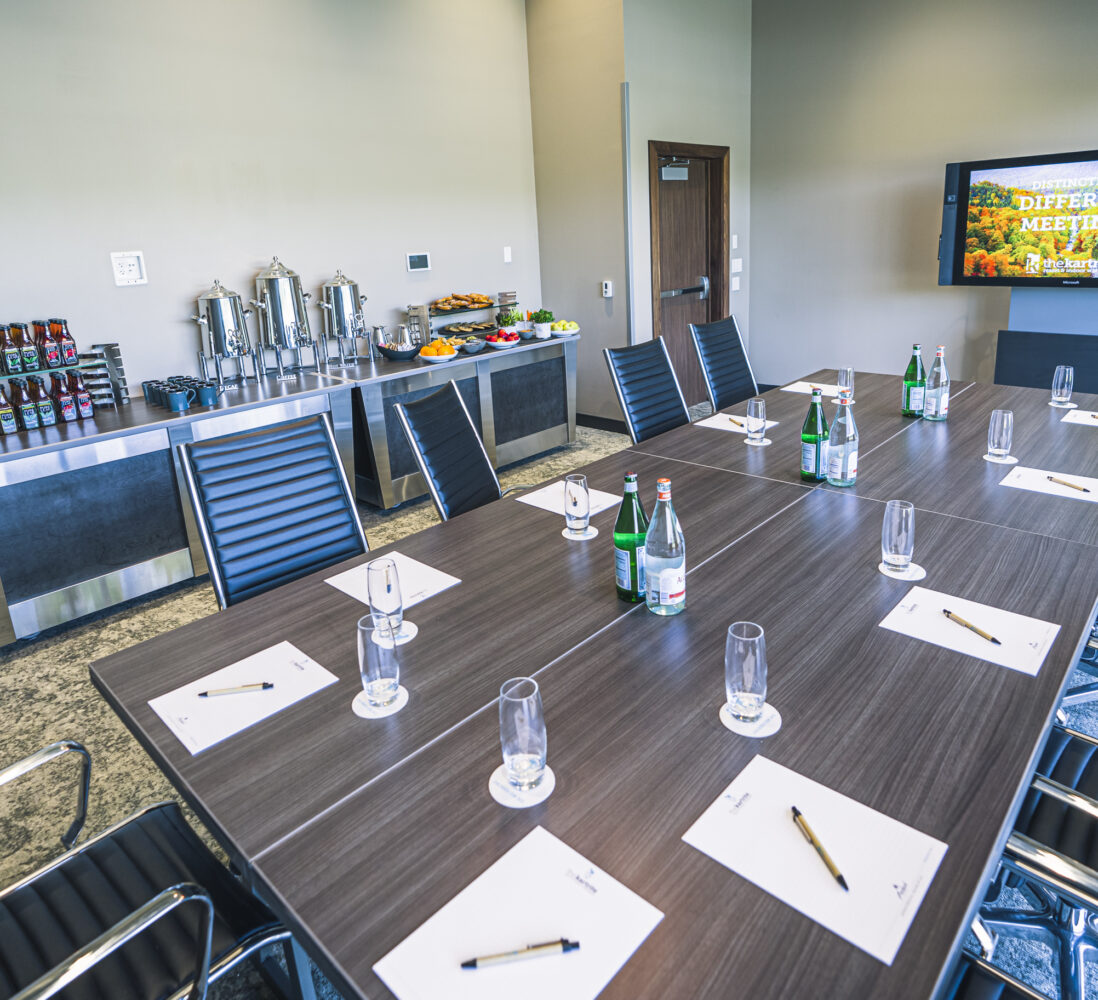 Modern conference room with a large table set for a meeting, drinks on the table, refreshments and coffee station along the wall, and a screen on the wall.
