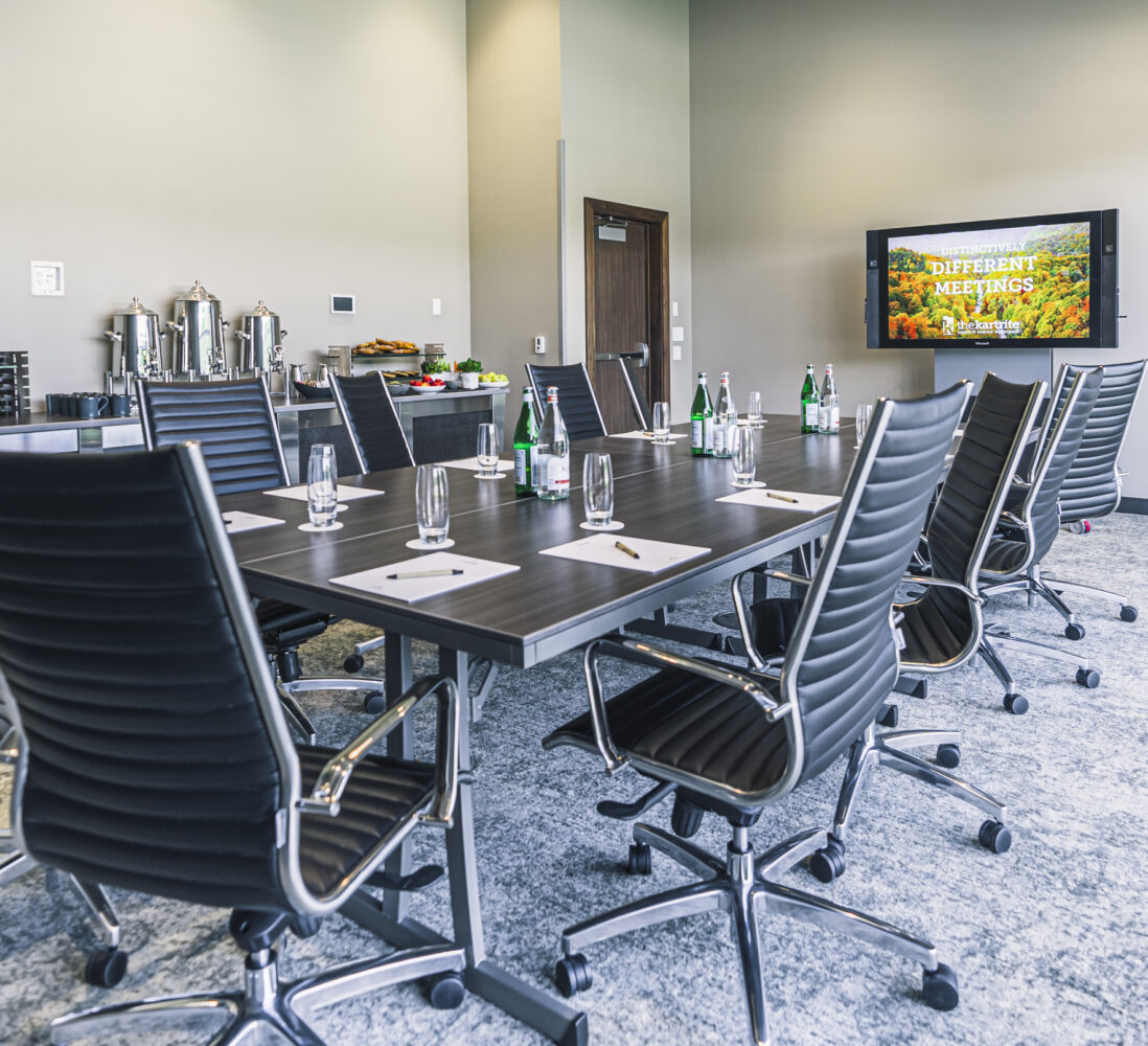 A modern conference room with a long table, black chairs, water bottles, notepads, and a TV displaying Confidently Host Meetings..
