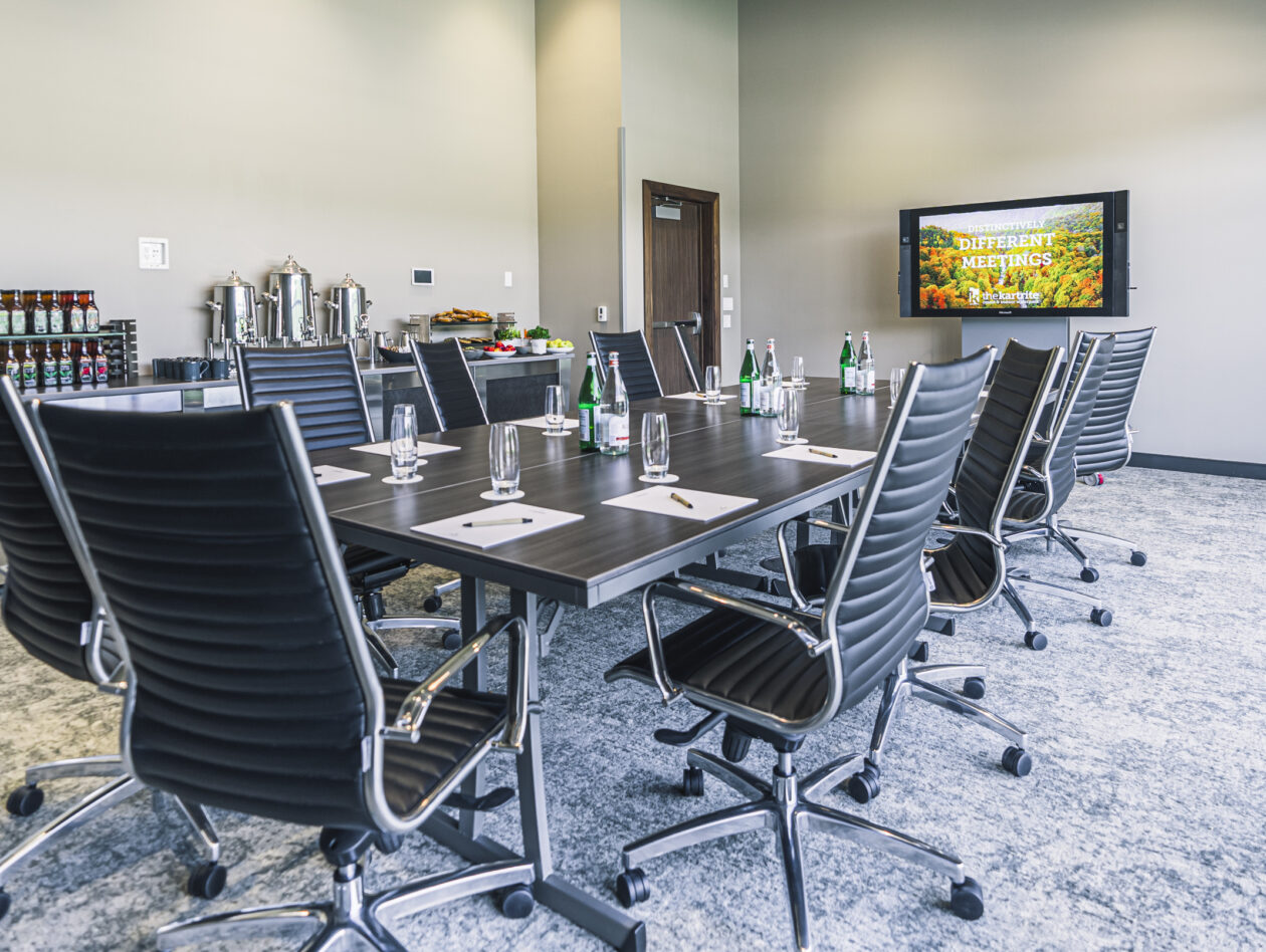 A modern conference room with a long table, black chairs, water bottles, notepads, and a TV displaying Confidently Host Meetings..