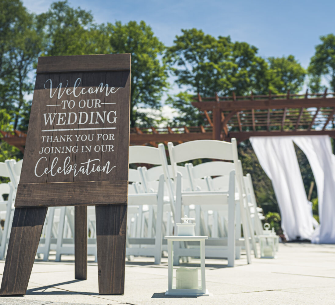 Outdoor wedding setup with white chairs, a wooden welcome sign, lanterns, and a wooden arch draped with white fabric under a blue sky.