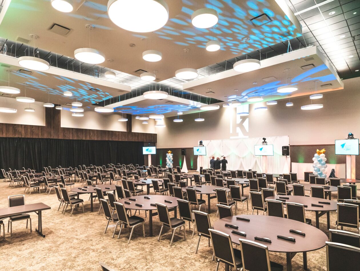 Spacious event hall with round tables, chairs, stage, large K on wall, balloon columns, and ceiling lights. Room is empty, set for an event.