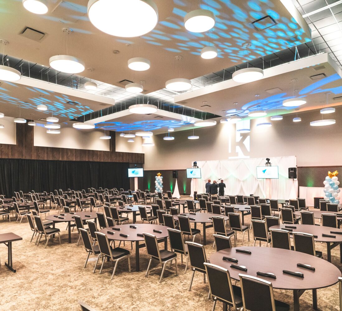 Spacious conference room with round tables, chairs, large stage, screens, and decorative lighting, prepared for an event or presentation.