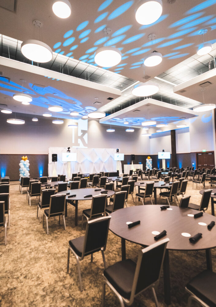 Large modern event hall with round tables, black chairs, blue lighting, and decorative patterns projected on the ceiling. Stage is set up at the front.