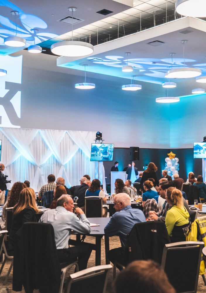 People sit at round tables in a large event room with blue lighting, watching a presentation on multiple screens.