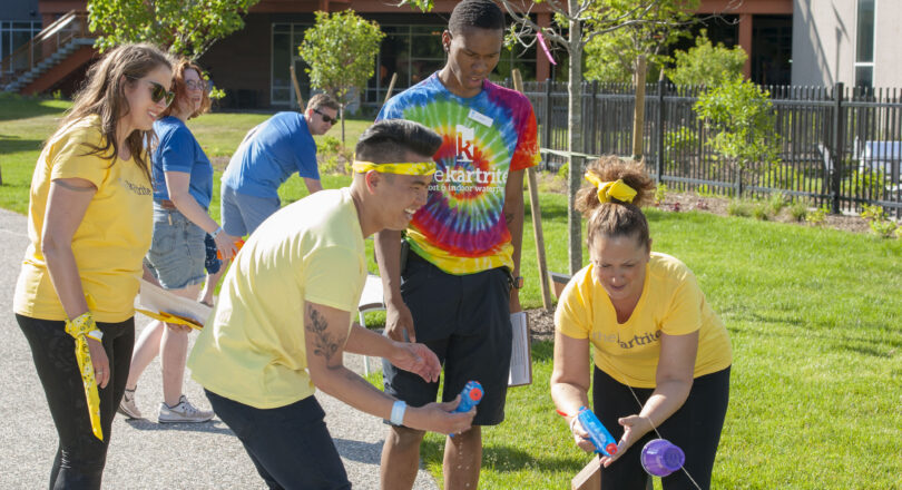 Four adults smile and play with water guns and cups on a sunny lawn, wearing bright shirts and headbands, with buildings in the background.
