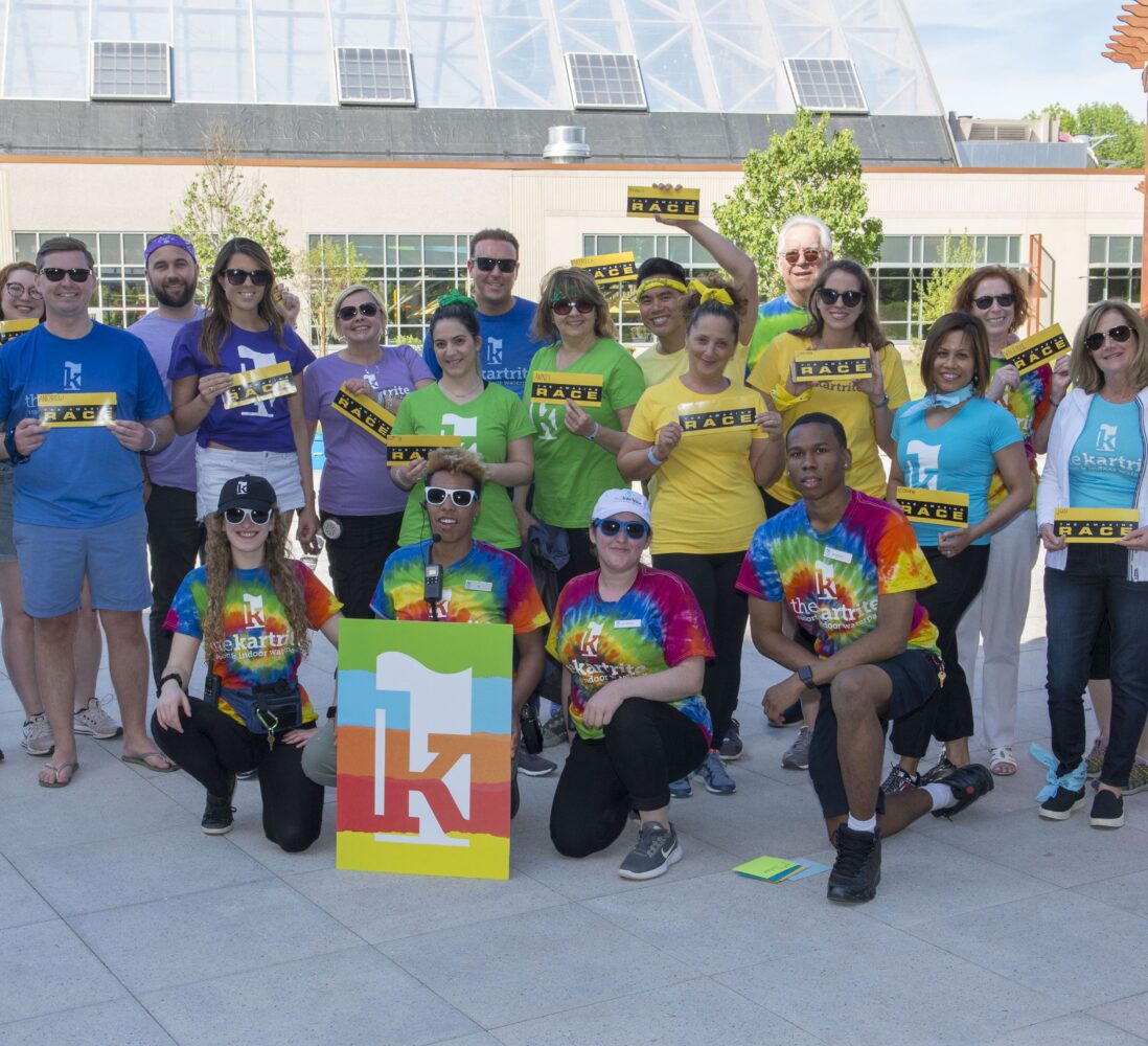 A group of adults wearing colorful shirts pose outdoors, holding signs and a large logo. Some are kneeling while others stand, all smiling at the camera.