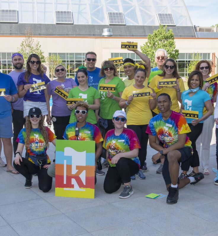 A group of adults wearing colorful shirts pose outdoors, holding signs and a large logo. Some are kneeling while others stand, all smiling at the camera.