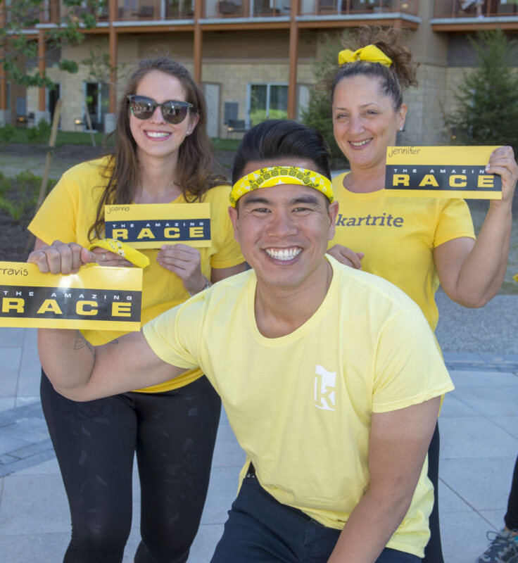 Three smiling people in yellow shirts hold The Amazing Race envelopes outdoors, wearing accessories and posing as a team.