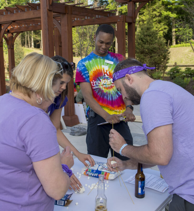 Four adults build a structure with marshmallows and pasta on a table outdoors; one wears a tie-dye shirt and others wear purple clothing and bandanas.