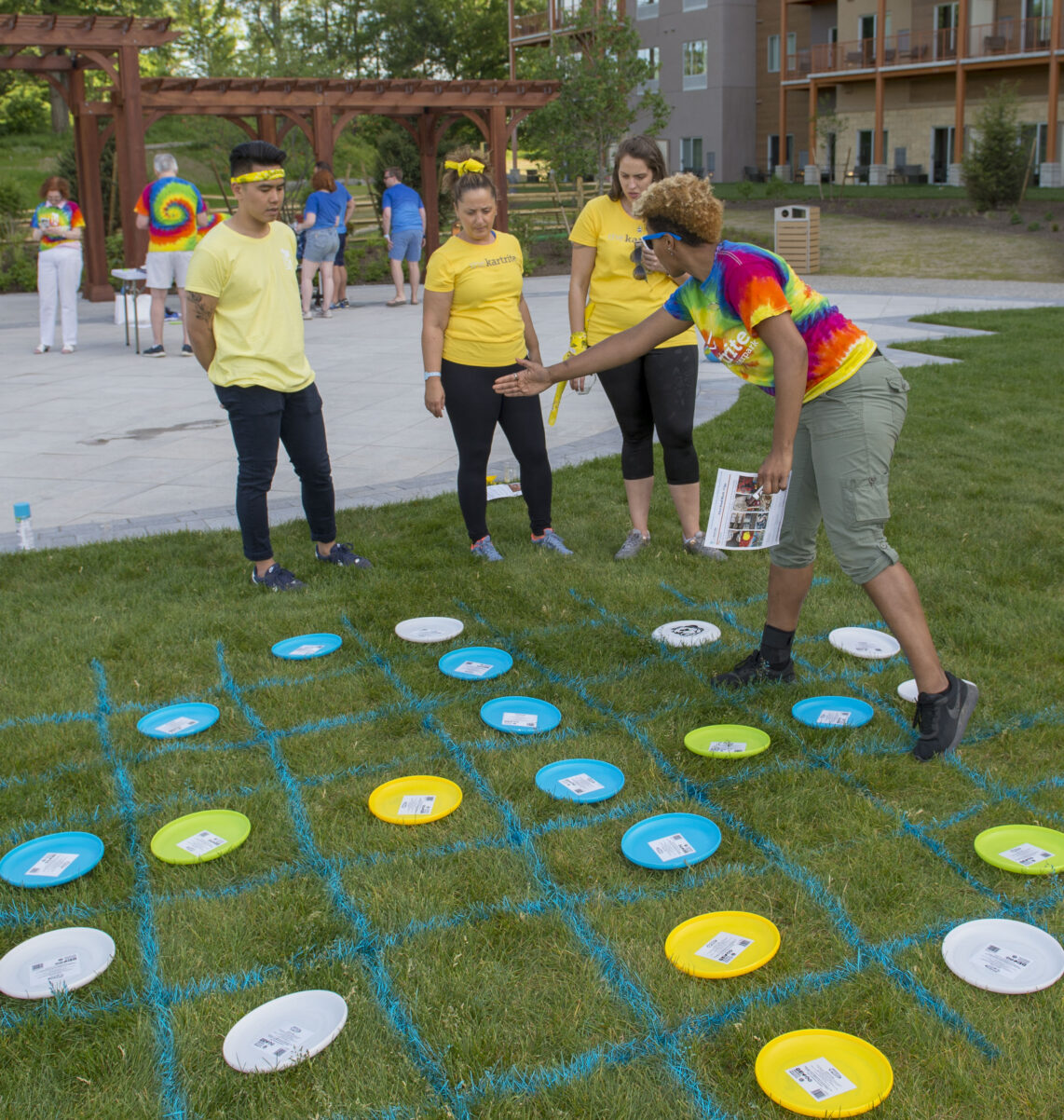 Four people play a large tic-tac-toe game outdoors using colored frisbees and blue grid lines painted on grass.
