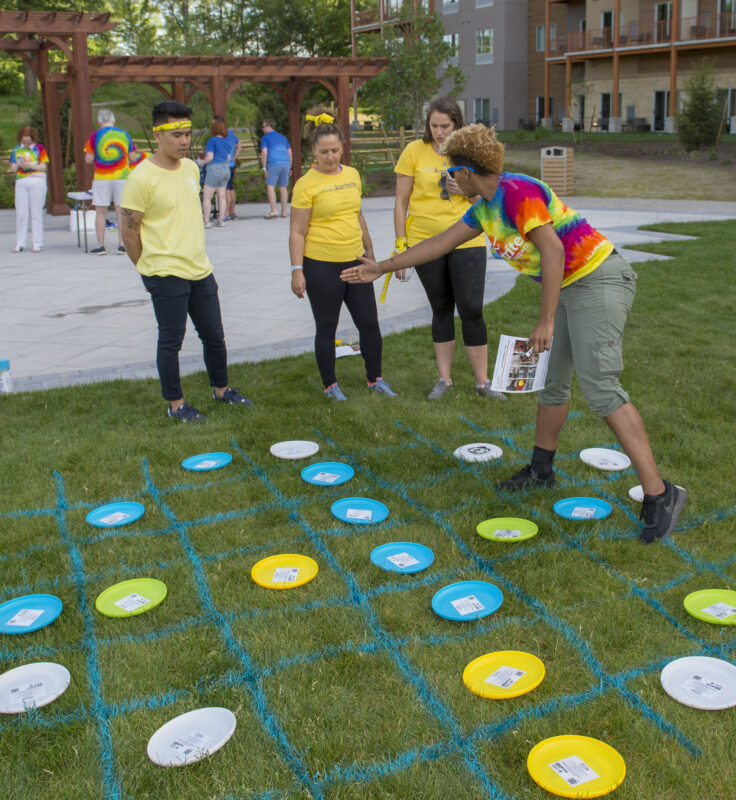 Four people play a large tic-tac-toe game outdoors using colored frisbees and blue grid lines painted on grass.