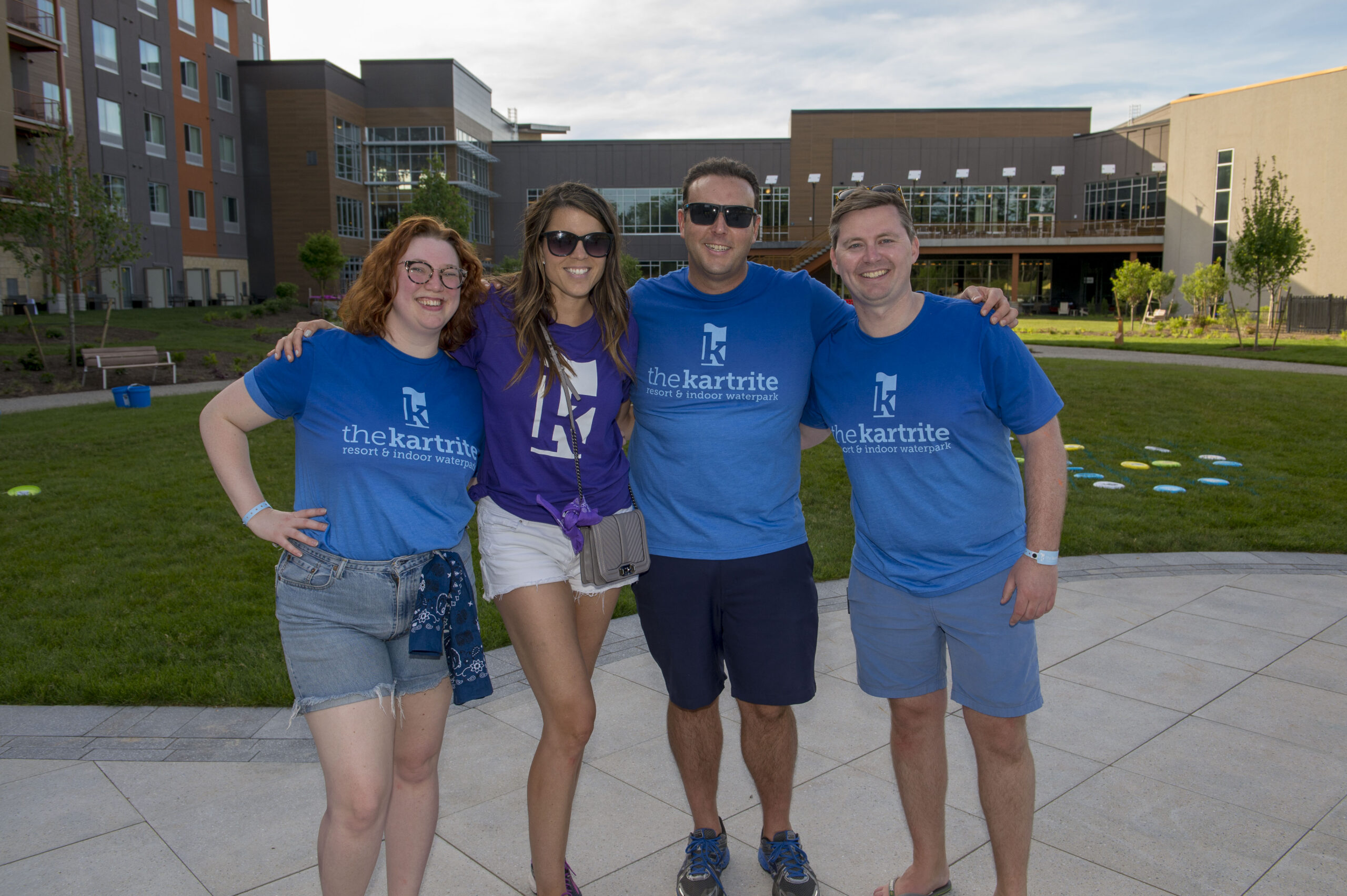 Four people stand smiling outdoors; two wear blue the kartrite shirts, one wears purple, and theyre in front of modern buildings and green grass.