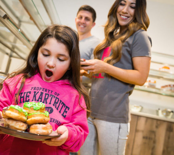 A young girl in a pink hoodie excitedly holds a plate of donuts, with two smiling adults standing behind her in a bakery.