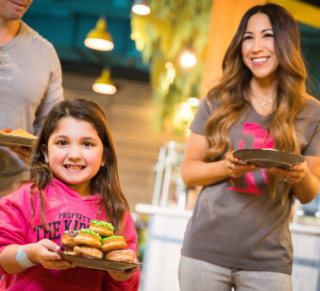 Smiling girl in a pink hoodie holds a plate of food, standing with two adults in a bright, casual dining setting.