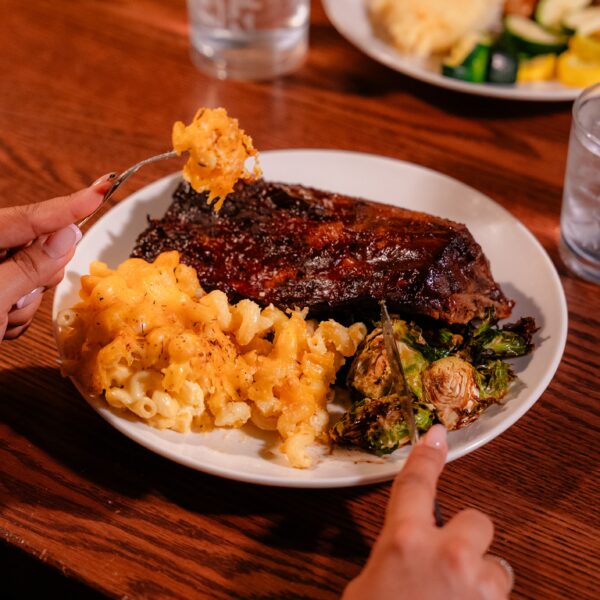 A person eating ribs, macaroni and cheese, and Brussels sprouts from a white plate, with a glass of water on a wooden table.