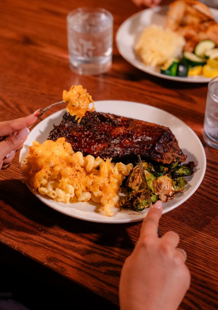 A person eating ribs, macaroni and cheese, and Brussels sprouts from a white plate, with a glass of water on a wooden table.