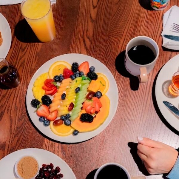 Colorful fruit platter, coffee, juice, granola bowl, pastries, pancakes, syrup, and two peoples hands on a wooden table set for breakfast.