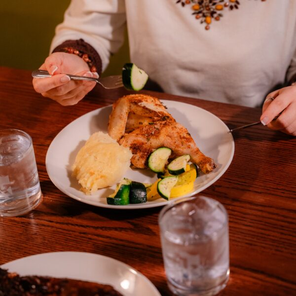 Person in a beaded top eating roasted chicken, mashed potatoes, and sautéed vegetables at a wooden table with glasses of water.
