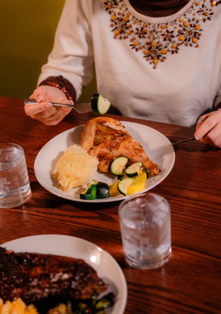 Person in a beaded top eating roasted chicken, mashed potatoes, and sautéed vegetables at a wooden table with glasses of water.