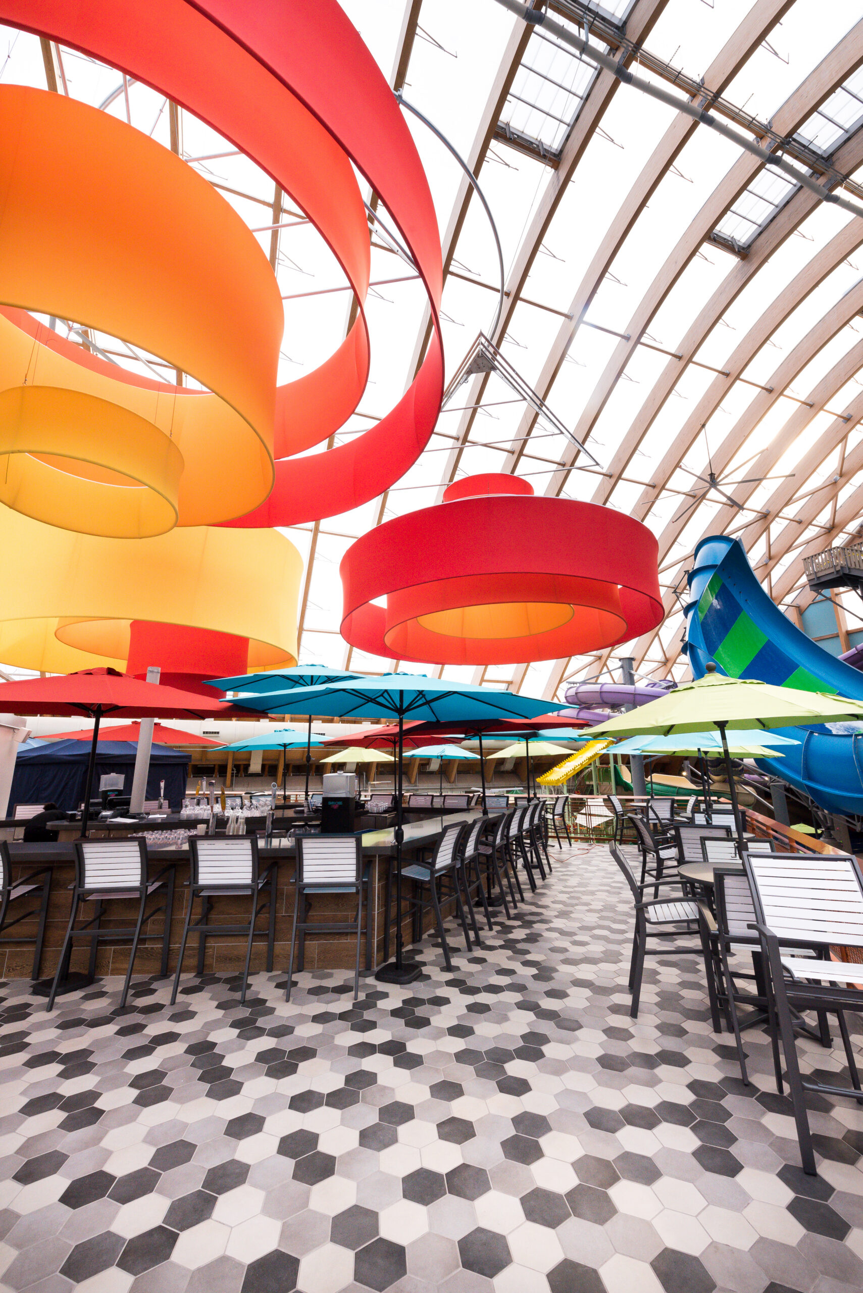 Colorful hanging spirals and umbrellas cover an empty outdoor dining area with black and white hexagonal tile flooring.
