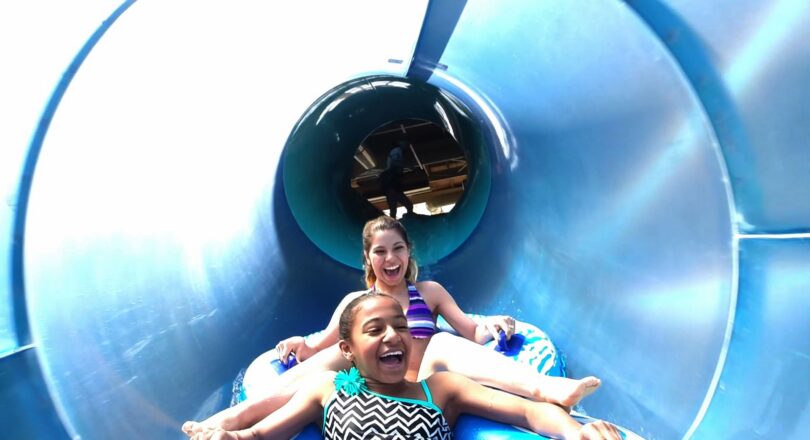 Two girls in swimsuits laugh while riding together on a blue inner tube down a twisting water slide at a water park.
