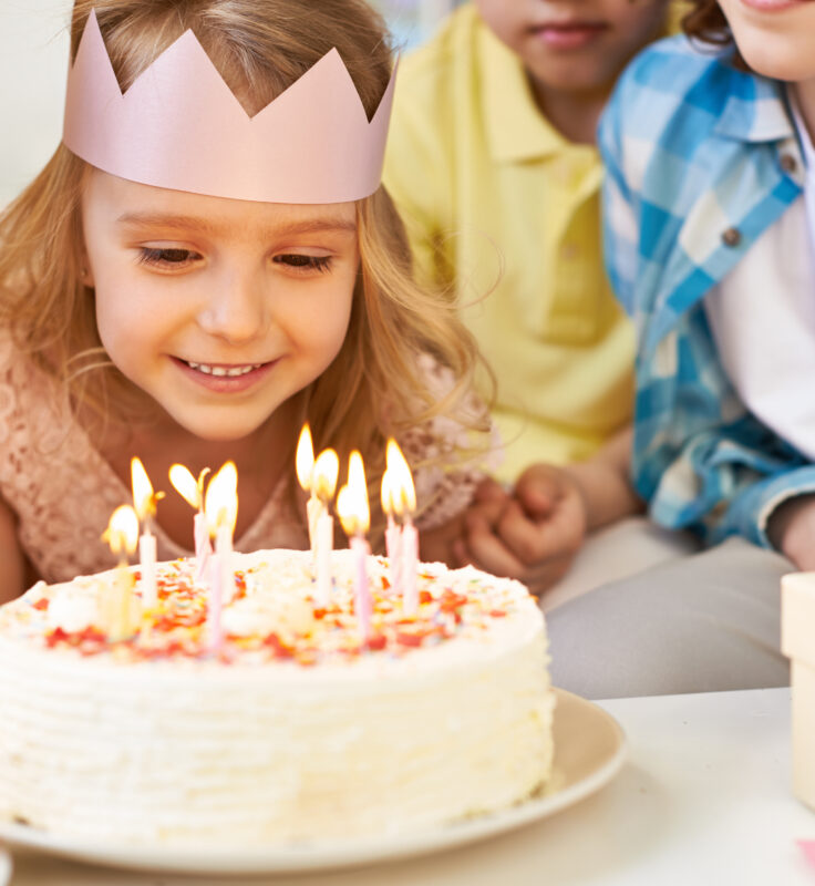 Smiling child wearing a paper crown prepares to blow out candles on a birthday cake, surrounded by friends.