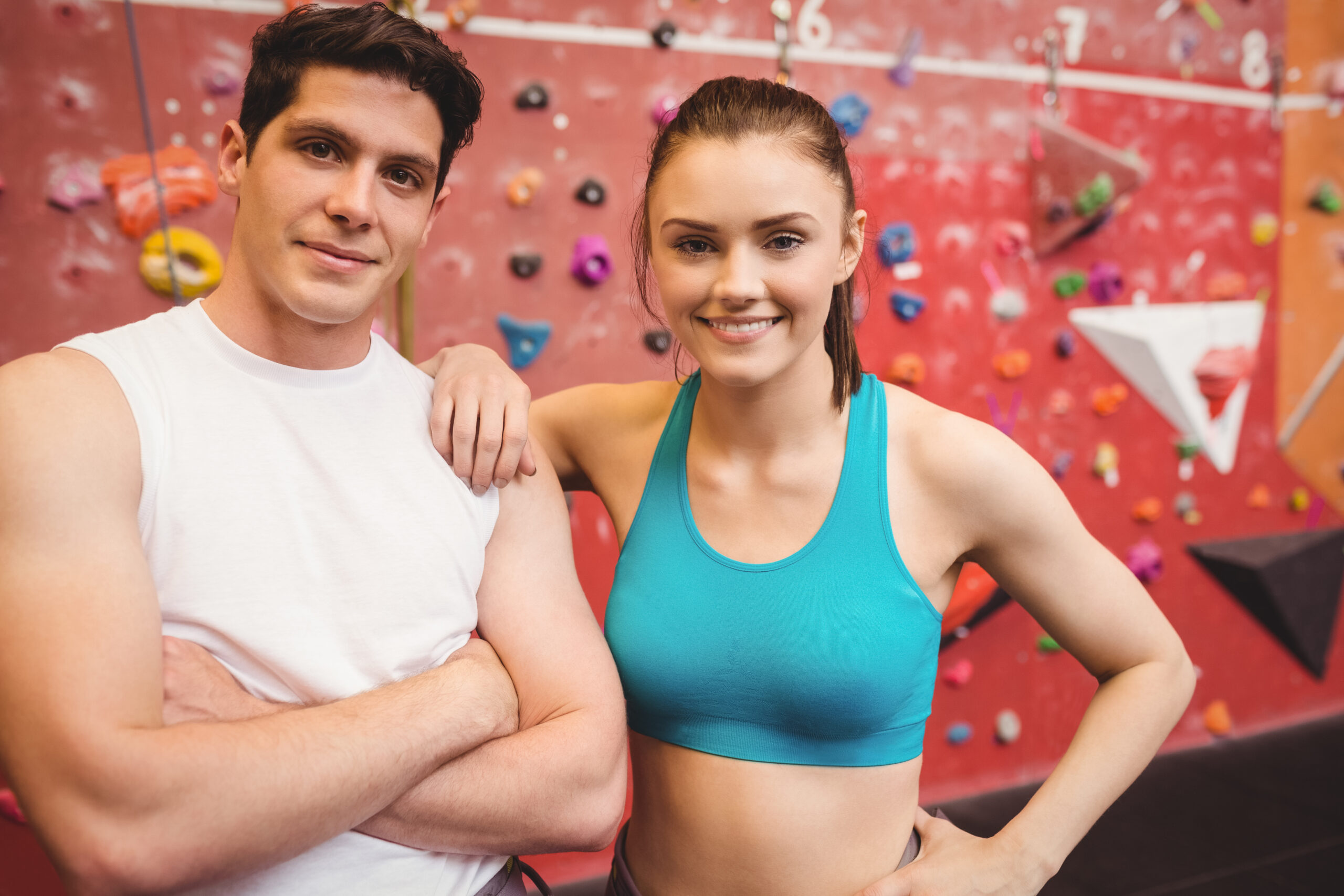 Two people in athletic wear smile at an indoor climbing gym, standing in front of a colorful climbing wall.