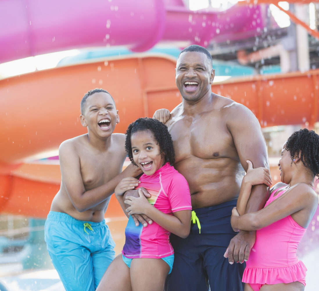 A man and three laughing kids stand in swimsuits at a water park, with colorful slides and splashing water in the background.