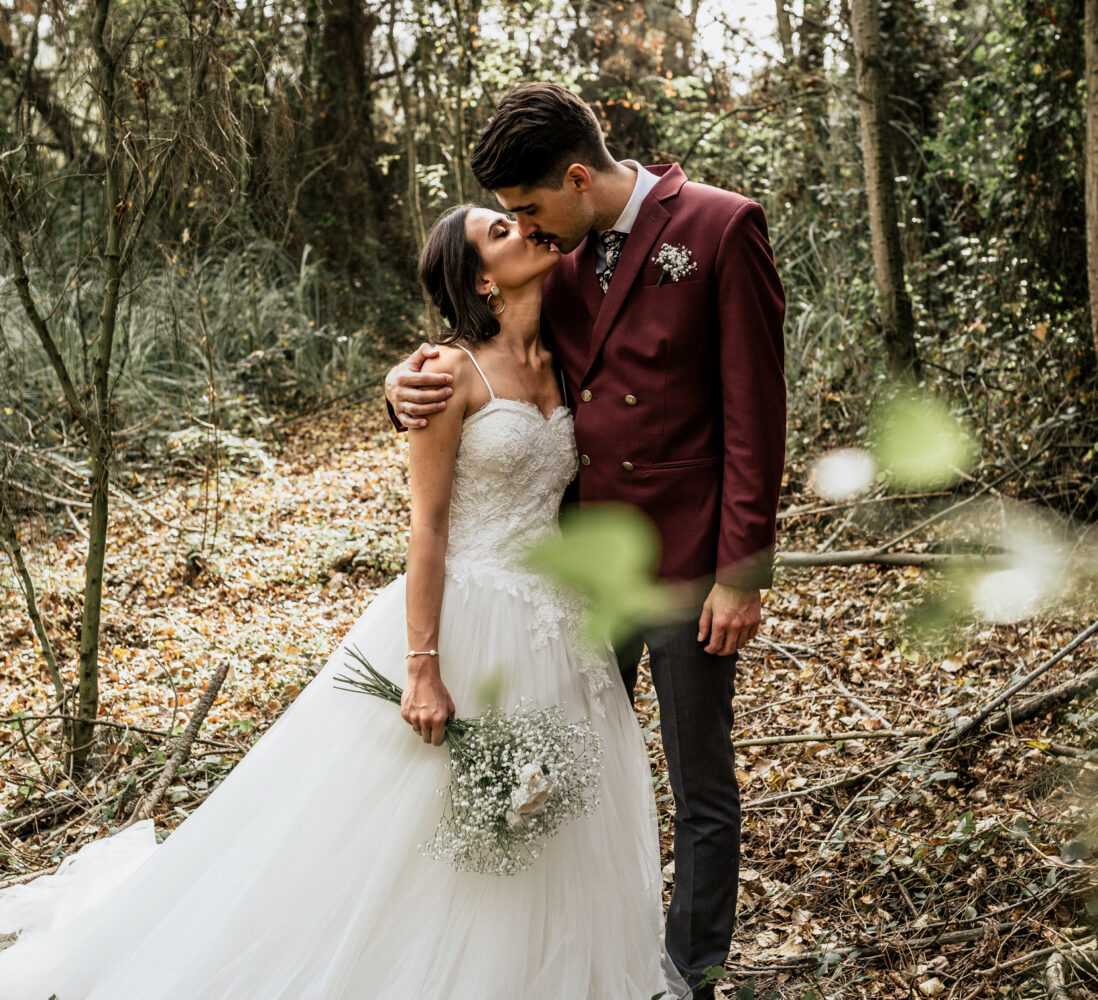A bride and groom stand in a forest, gently kissing, with the bride in a white dress holding flowers and the groom in a burgundy suit.