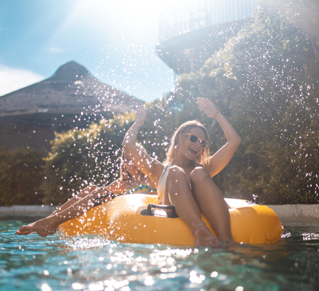 Two people relax on a yellow float in a sunlit pool, splashing water and smiling, surrounded by greenery and buildings in the background.