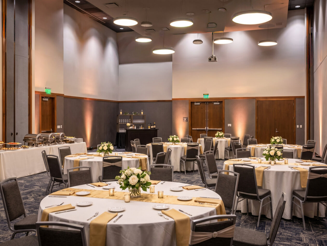 Elegant banquet hall with round tables set for an event, white tablecloths, gold runners, floral centerpieces, and buffet tables along the wall.