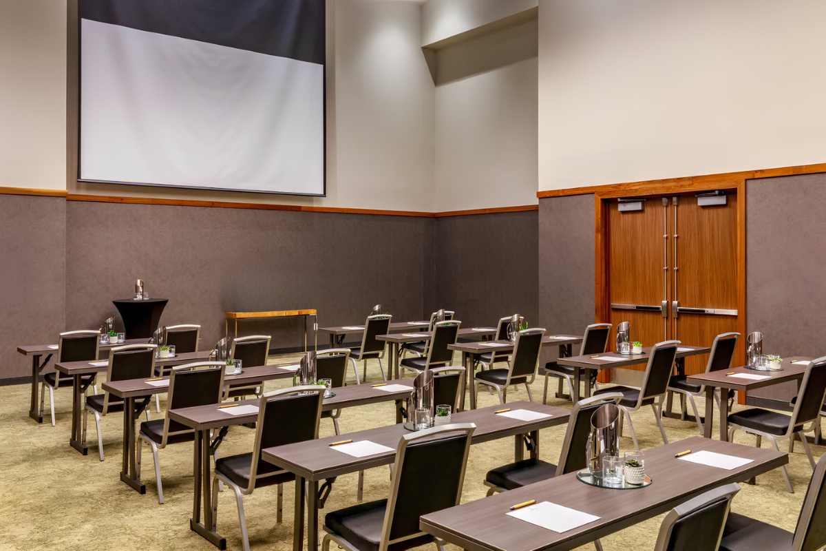 Conference room with rows of tables and chairs facing a large screen, each table set with paper, pens, water glasses, and pitchers.