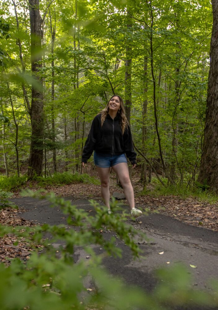 A person in a black hoodie and shorts walks on a forest path, smiling and surrounded by green trees and foliage.