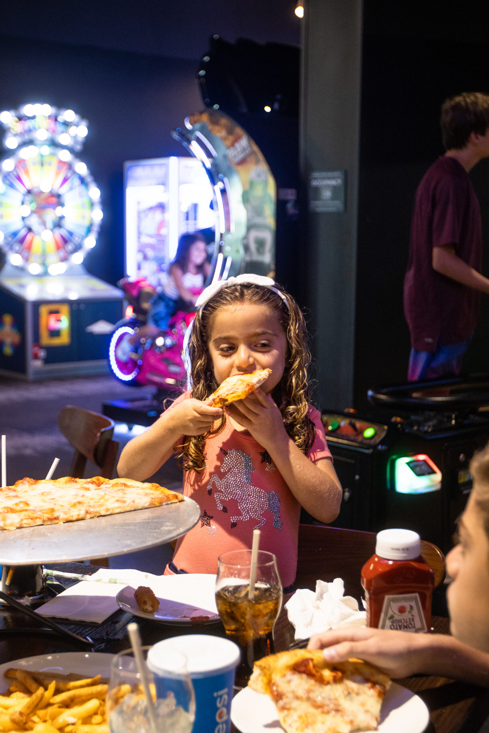 Young girl with curly hair eats pizza at an arcade, with game machines and other people in the colorful, lively background.