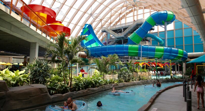 People float on inner tubes along a lazy river inside an indoor water park with large, twisting water slides and tropical plants.