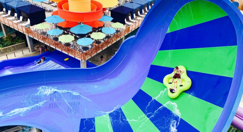 A person rides a green inflatable tube down a bright blue and green indoor water slide near colorful orange spiral slides and poolside seating.