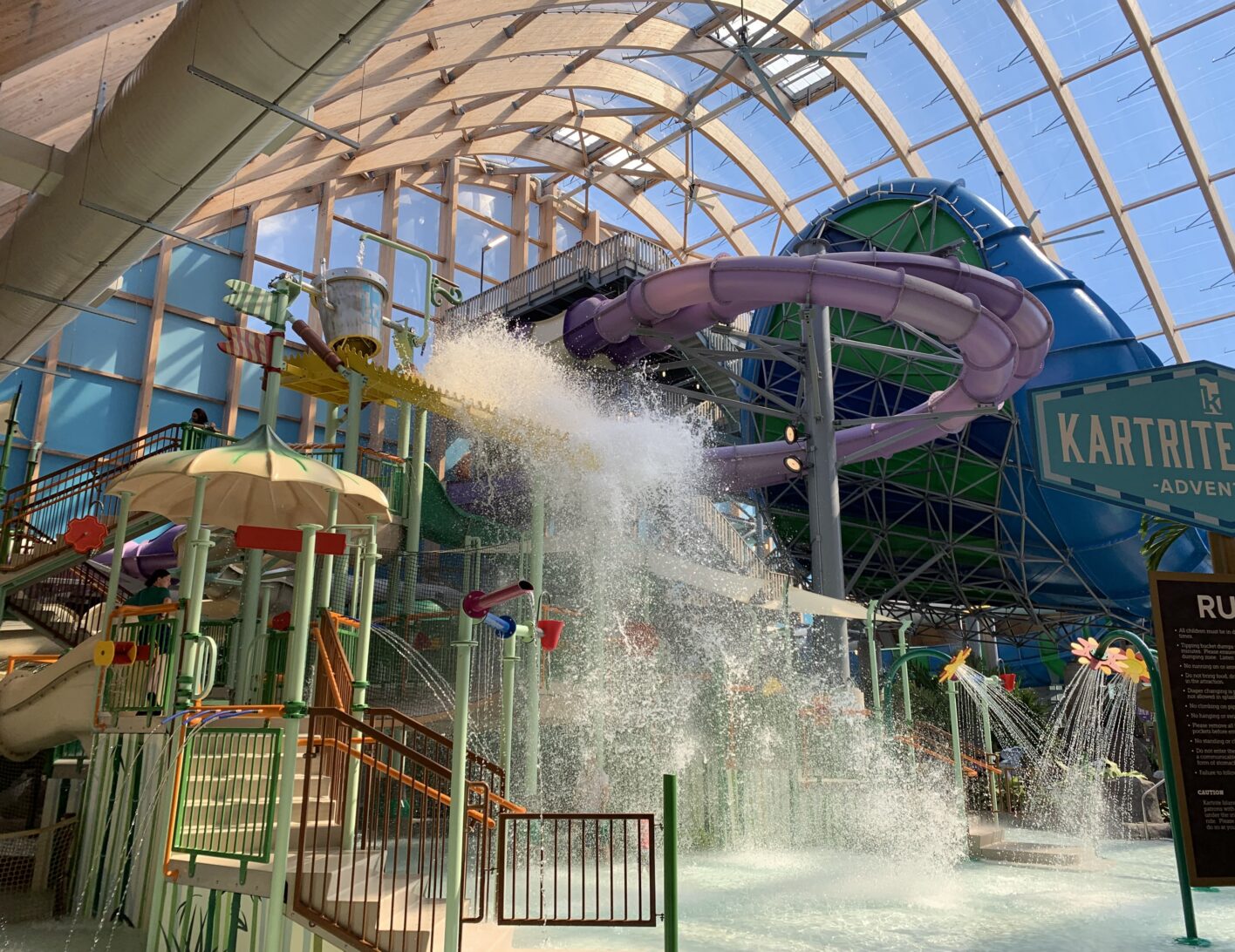 Large indoor waterpark with a giant tipping bucket spilling water, purple waterslides, and bright sunlight streaming through the glass roof.