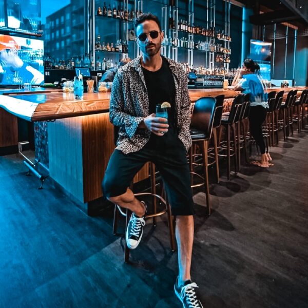 Man in sunglasses and patterned shirt poses with a drink at a stylish bar, one leg up on a stool. Shelves of bottles and patrons visible in background.